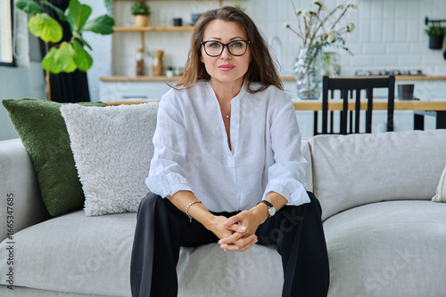 Fotografie Successful beautiful mature woman looking at camera sitting at home on sofa