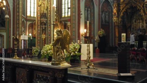 Wallpaper Mural Blurred view of a catholic church interior with a golden angel statue, flowers, and blurred figures in background, creating a serene, sacred atmosphere. Torontodigital.ca