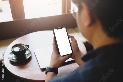 Over the shoulder view of a man holding a smartphone with a blank white screen, sitting in a cafe with coffee. Ideal for app or website mockups.