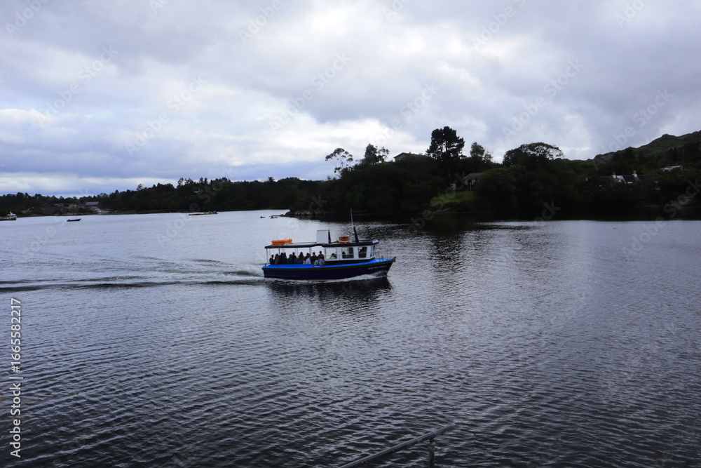 Fototapeta premium Scenic Boat Ride on Calm Lake with Tourists Enjoying Nature and Tranquil Waters, Surrounded by Green Hills, Forest, and Cloudy Sky – Perfect for Travel, Adventure, and Relaxation Concepts