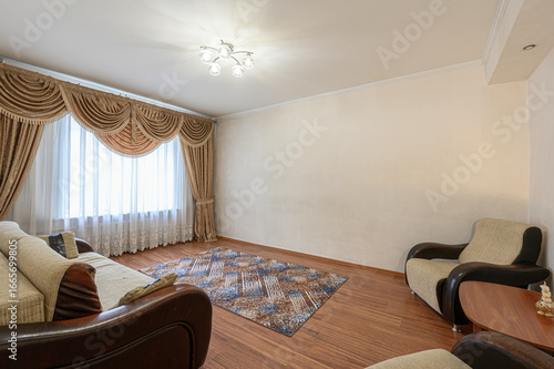 Cozy living room with brown wooden floor, beige walls, ornate curtains, a patterned rug, and a sofa armchairs