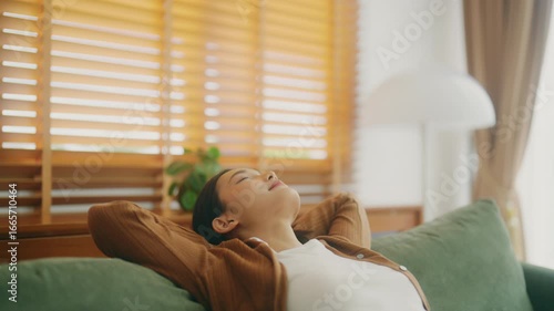 Relaxed young Asian woman enjoying rest on comfortable sofa, calm attractive girl relaxing on couch, breathing fresh air with eyes closed, meditating at home