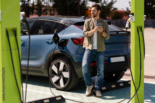 Canvas Print Man using smartphone while charging electric car at charging station