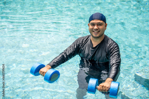 Tableau sur toile A smiling person in a swim cap and rash guard is submerged in a pool, holding blue aquatic dumbbells