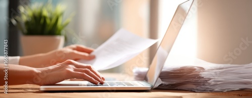 The laptop with hands typing, papers stacked nearby, sunlight and office plant