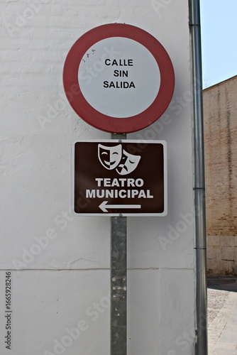 A street sign on a white wall displaying a “dead-end street” warning above a brown municipal theater directional sign with comedy and tragedy masks in spanish town