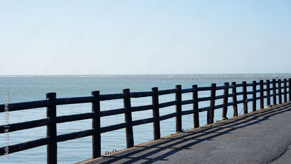 Fototapeta premium A coastal walking path with a black wooden fence casting shadows on the ground, overlooking calm ocean water. Wind turbines are visible on the distant horizon.
