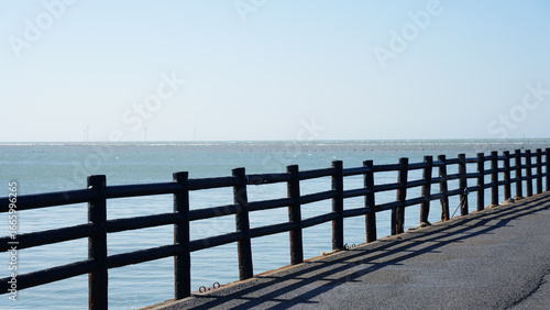 A coastal walking path with a black wooden fence casting shadows on the ground, overlooking calm ocean water. Wind turbines are visible on the distant horizon.