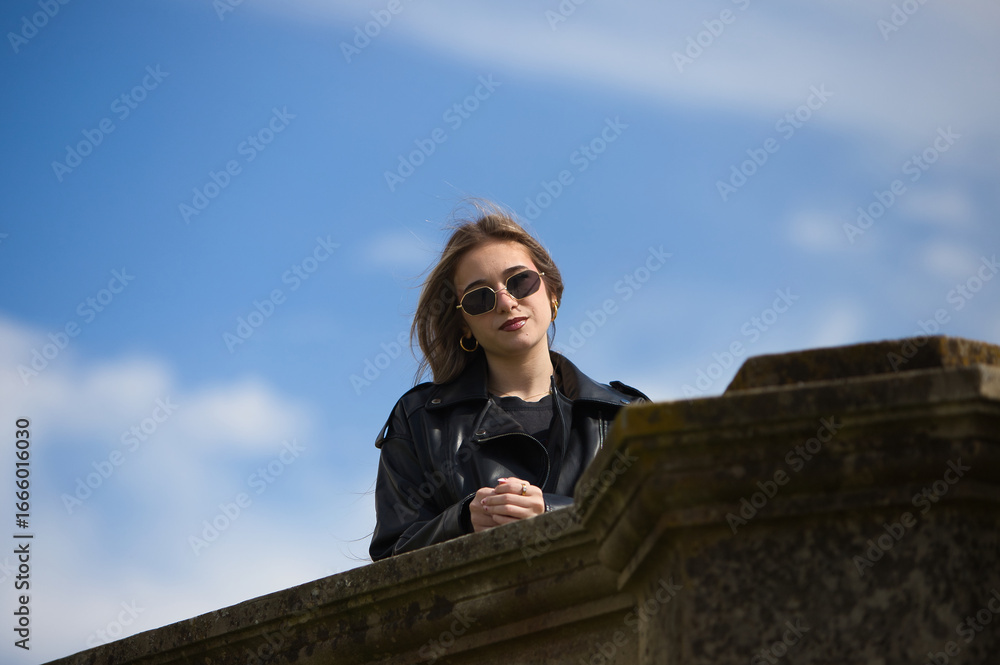 Fototapeta premium pretty young blonde woman with sunglasses, black leather jacket, boots and short skirt leaning against the wall with cool attitude. The woman is smiling and happy. In the background the blue sky.