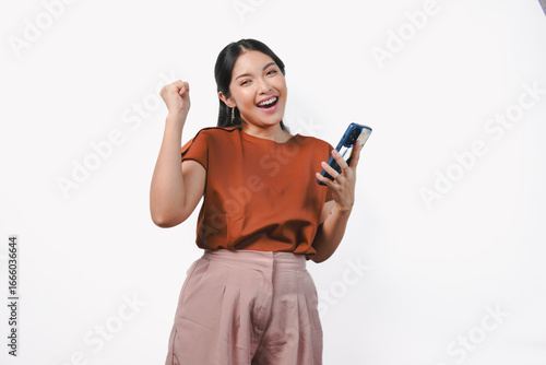 Excited young Asian woman raising her fists with happy successful expression while holding phone.