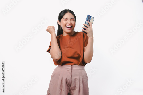 Excited young Asian woman raising her fists with happy successful expression while holding phone.
