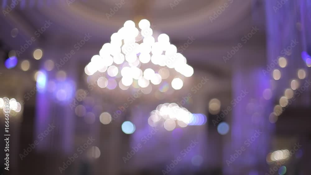 A grand, ornate crystal chandelier hangs from a beautifully decorated ceiling, reflecting light and adding a touch of luxury and elegance to the venue.