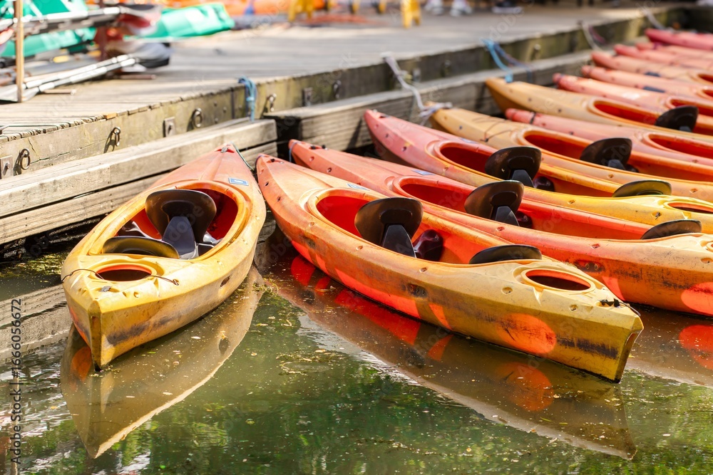 Obraz premium Colorful kayaks in vibrant shades of orange and yellow are lined up along a wooden dock, reflecting in the calm water, creating a serene outdoor adventure scene
