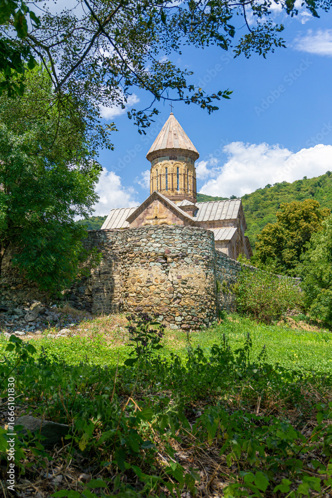 Obraz premium The outside of a monastery with a church and high stone walls. Green grass and trees. Hills, sky with clouds in the background. Pitareti monastery. Georgia