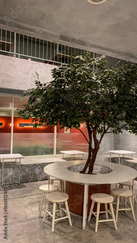 Modern cafe interior with a tree centerpiece, round table, and wooden stools, combining natural and minimalist industrial design.