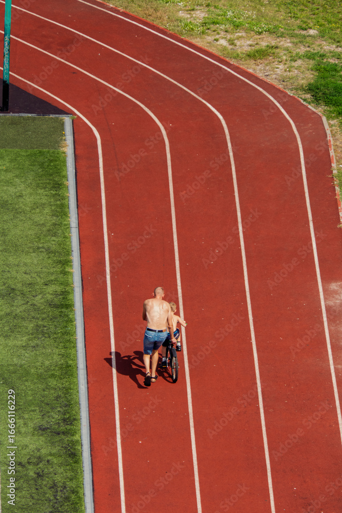 Obraz premium Father teaching his son to ride a bicycle on a running track. A touching moment of family bonding and learning, viewed from above. Childhood development and outdoor activity.