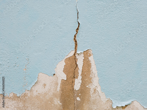 Close-up light blue painted wall with large, peeling sections revealing gray, rough concrete underneath. Old paint is flaking and cracked