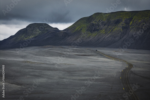 Aerial view of a stark, grey road winding through an ashen landscape, contrasted by dark, moss-streaked mountains under a brooding sky, highlands, Iceland.