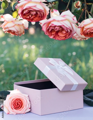 A light mauve gift box, open and ready to receive a treasured gift, rests on a table, surrounded by beautiful, vibrant, and elegant roses in a lush garden setting.