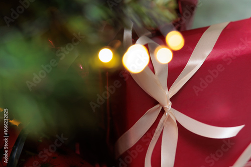 immagine in tema natalizio, con pacco regalo infiocchettato di colore rosso sullo sfondo, con rami di un albero di natale e luci sfuocato in primo piano