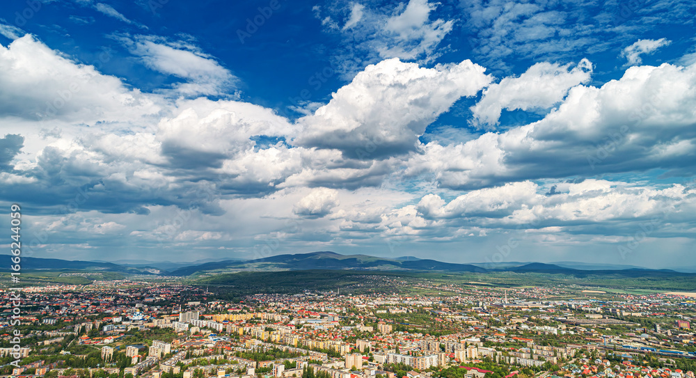Fototapeta premium An aerial panoramic view of a city surrounded by mountains and greenery under a dramatic cloudy sky.