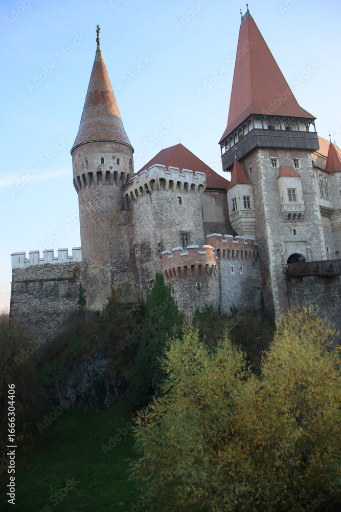 Fototapeta premium A grand medieval castle with tall red-roofed towers, stone walls, and battlements, rising above trees and rocky cliffs, showcasing Gothic architecture and historic charm.