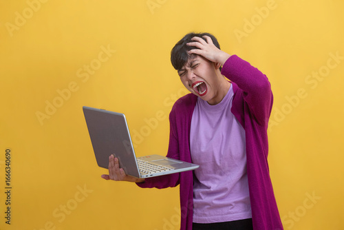 Frustrated woman shouting while holding a laptop in one hand and clutching her head with the other, standing against a bright yellow background. Concept of tech, work stress and digital frustration.