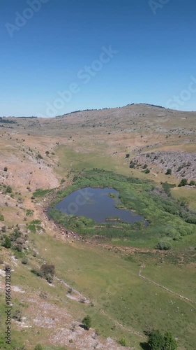 Drone aerial footage of a small mountain lake surrounded by rocky hills and green vegetation, representing untouched nature and scenic landscape.