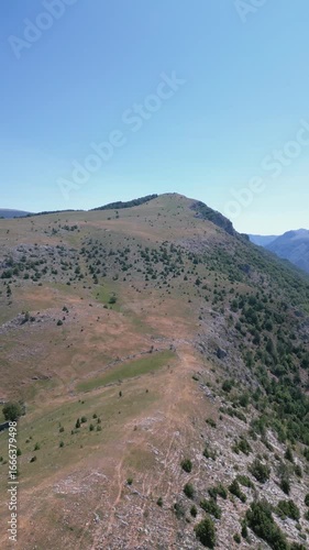 Aerial drone view of a rocky mountain ridge with scattered vegetation and clear blue sky, representing natural landscape and wilderness.