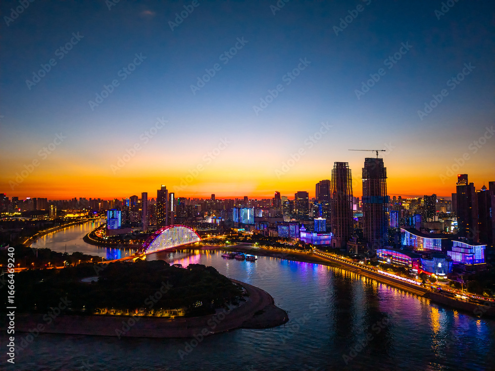 Fototapeta premium Wuhan city in China presents a radiant evening skyline along the Yangtze River, with colorful lights illuminating modern buildings and a bright bridge reflecting on the water.