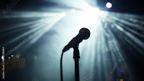 A lone microphone stands ready on a dimly lit stage, bathed in the dramatic glow of bright spotlights and atmospheric haze, awaiting the next vocal performance