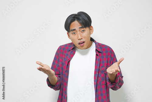 Young man with surprised expression, gesturing with open hands, wearing a red and blue plaid shirt over a white t-shirt.