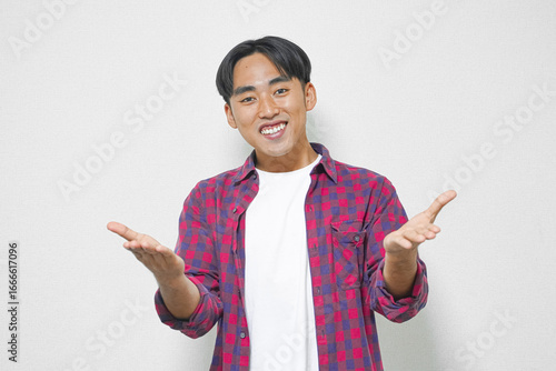 Young Asian man smiling and gesturing with open hands, wearing a red and purple plaid shirt over a white t-shirt.