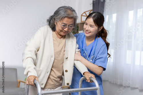 Asian Elderly woman using walker with support from smiling female nurse in blue uniform inside bright room with window and curtain