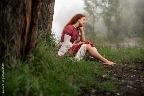 Redhead woman in flowing dress sits pensively against a tree in misty forest setting.