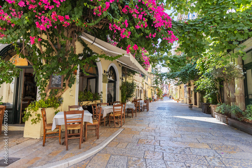 Fototapeta Naklejka Na Ścianę i Meble -  Picturesque Mediterranean street lined with vibrant bougainvillea flowers, outdoor dining tables, and charming architecture. Scene captures the essence of a lively yet peaceful coastal town. Corfu.