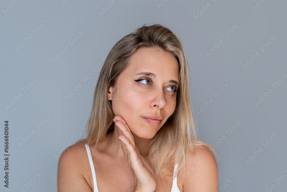Young blonde woman gently touching her face, looking away thoughtfully. Skincare, reflection, natural beauty, facial contour, mindful moment on neutral background Young blonde woman gently touching her face, looking away thoughtfully. Skincare, reflection, natural beauty, facial contour, mindful moment on neutral background