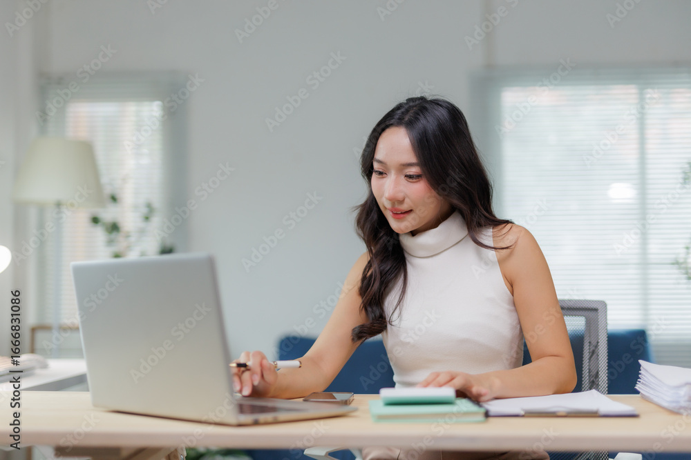 Asian businesswoman working with laptop computer, holding pen and looking at screen, sitting at desk in modern office with documents and notebooks Asian businesswoman working with laptop computer, holding pen and looking at screen, sitting at desk in modern office with documents and notebooks