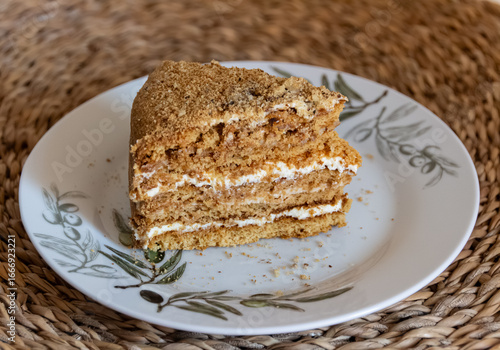 Piece of cake (medovnik) is on a plate. Layered cake, with crumbs around it. Served on a white plate on a wooden place mat. Closeup photograph from above. Version 1.