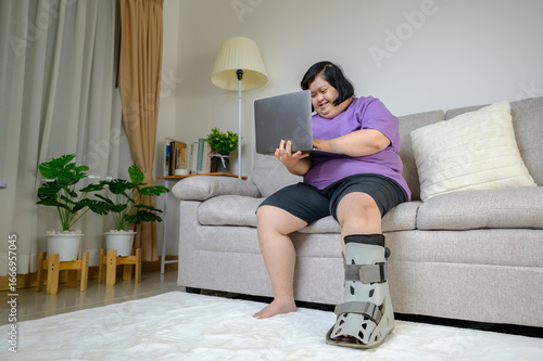 Asian woman wearing a leg brace sitting on sofa, holding laptop and smiling with excitement at home. Concept of disability, rehabilitation, healthcare, diversity, inclusion, and positive lifestyle.