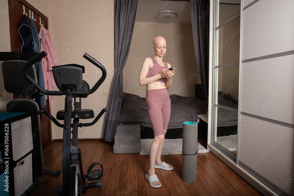 Bald woman with cancer in sportswear using smartphone before home fitness workout Bald woman with cancer in sportswear using smartphone before home fitness workout