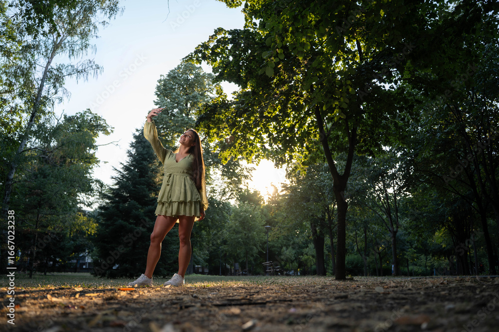 Fototapeta premium Happy beautiful young woman taking smiling selfie in park at golden hour sunset.