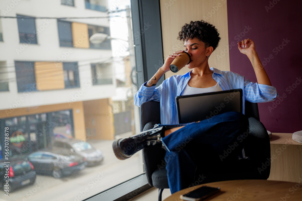 Obraz premium Young creative freelancer woman is drinking coffee and working on a laptop, enjoying her morning routine in a modern office space with a view of the city