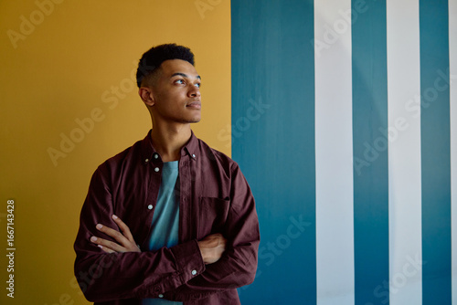 Thoughtful young businessman in smart casual standing arms crossed against colorful wall in office 