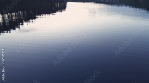 A fast, dynamic low-angle flight over the calm water surface of a lake, capturing stunning reflections of the surrounding forest and sky at sunset
