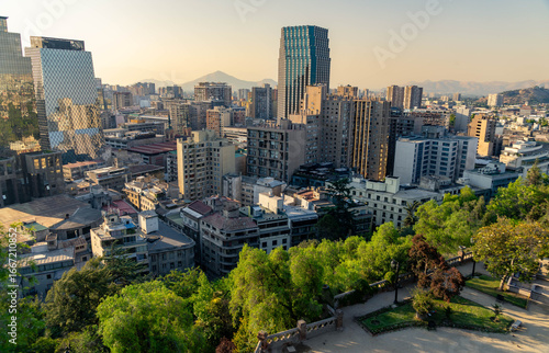 Skyline of Santiago, Chile at sunset with modern skyscrapers, historic buildings, and the Andes mountains in the distance, viewed from Cerro Santa Lucía with lush greenery in the foreground.