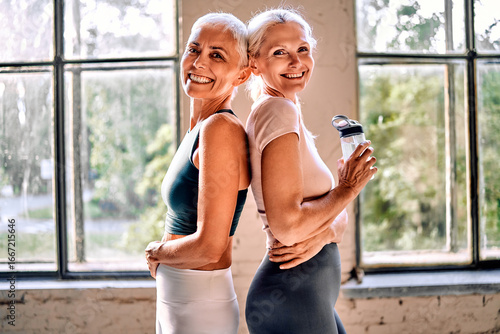 Two beautiful older women with grey hair wearing sportswear standing in a fitness gym. Motivation, physical activity, and a healthy lifestyle.