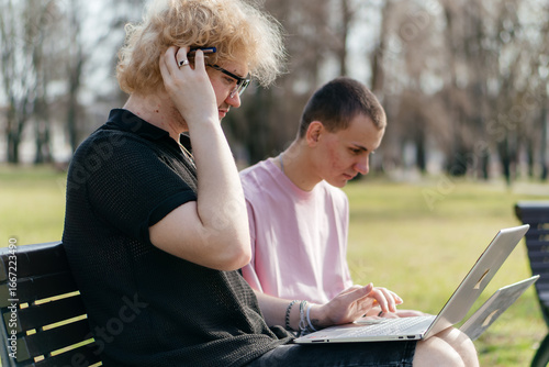 Three cheerful students study and work together outdoors on a sunny day, using laptops and notebooks while sitting on a park bench