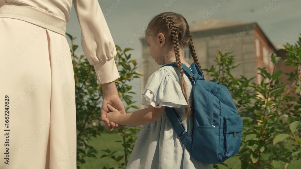Fototapeta premium mom leads her little daughter with a backpack to school, escort child to preschool education, walk home through schoolyard, first grade student, happy family, kid with school bag by hand with mother.