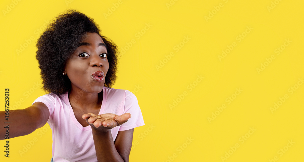 Joyful african american lady taking selfie, blowing air kiss to camera, having fun on yellow studio background, free space. Lovely young woman posing to mobile camera and making photo of herself Joyful african american lady taking selfie, blowing air kiss to camera, having fun on yellow studio background, free space. Lovely young woman posing to mobile camera and making photo of herself
